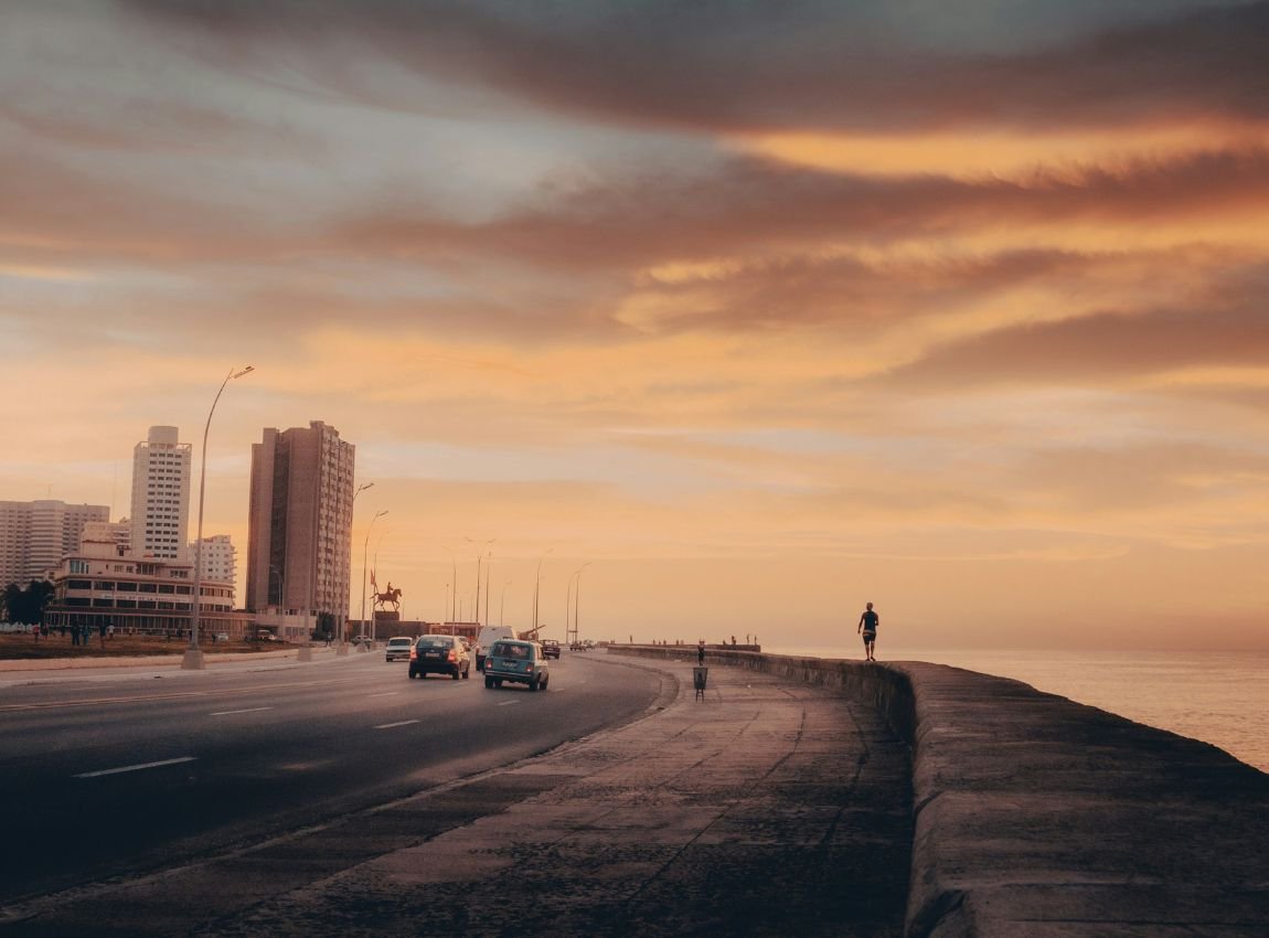 2: Vista del icónico Malecón de La Habana al atardecer. Un corredor junto al mar con el cielo teñido de tonos dorados y rosados, donde autos y peatones disfrutan de la brisa caribeña.