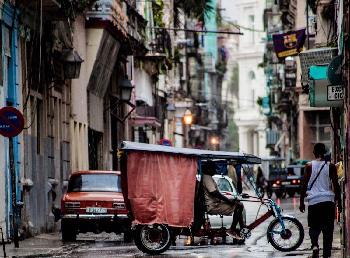 Una vista de una calle mojada en La Habana, Cuba, con un bicitaxi en primer plano, un coche rojo y personas caminando. Los edificios tienen balcones antiguos