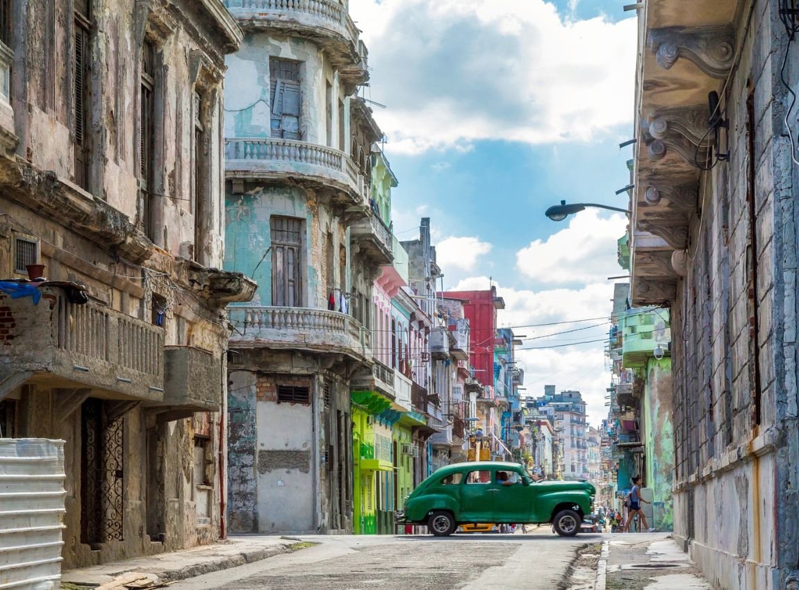 Calle en La Habana, Cuba, con un coche verde antiguo pasando entre edificios coloridos y balcones deteriorados bajo un cielo azul con nubes.